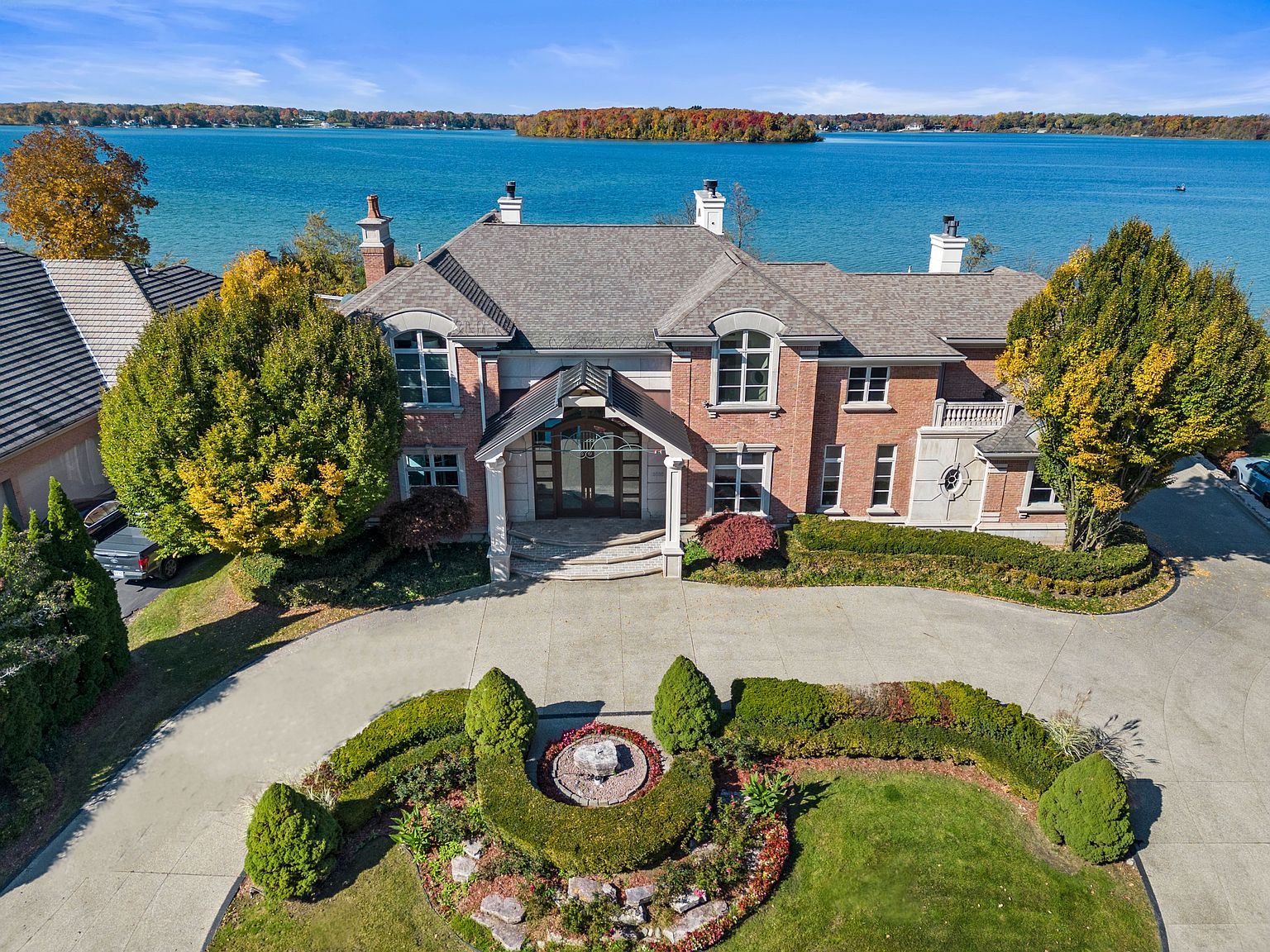 This aerial shot showcases a grand brick house with a meticulously landscaped circular driveway and garden. The house features a complex roofline with multiple chimneys and dormers, suggesting a luxurious and spacious interior. The property is situated on a waterfront, offering stunning views of the lake and distant wooded islands, enhancing its appeal as a high-end real estate offering.