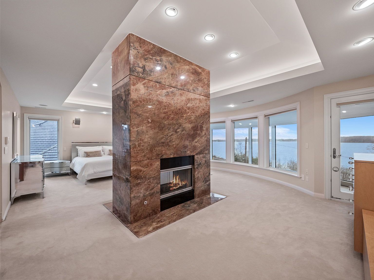This is a primary bedroom featuring a unique central fireplace clad in marble, dividing the room into sleeping and sitting areas. The bedroom boasts large windows with water views, recessed lighting, and neutral carpeting. A mirrored dresser adds a touch of glamour, and the overall impression is luxurious and serene.
