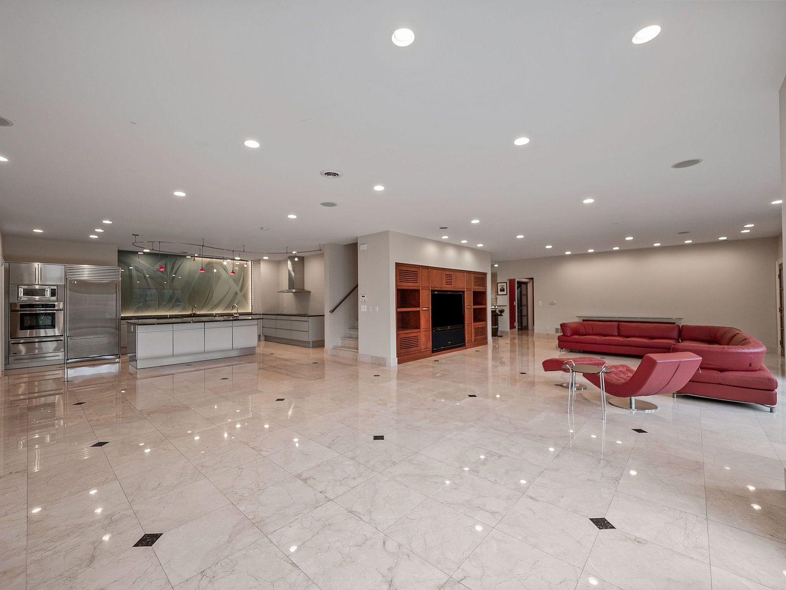 This expansive living room seamlessly integrates with a modern kitchen, featuring a sleek stainless steel refrigerator and oven. The open floor plan boasts a glossy tiled floor, recessed lighting, and a built-in entertainment center with wooden cabinetry. A red leather sofa and chair set adds a pop of color to the neutral palette, creating a luxurious and inviting atmosphere.