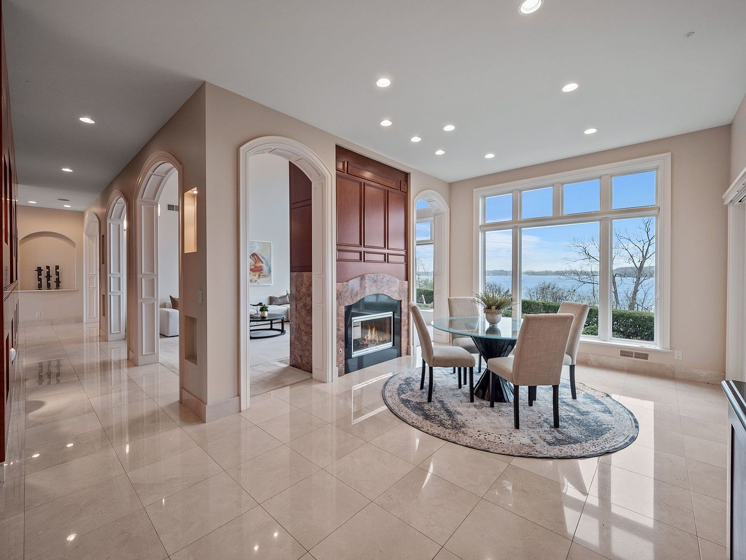 This interior shot showcases a bright and elegant dining area with a large window offering a scenic view of the water. The room features a round glass table with four chairs, set on a patterned rug, and a fireplace with dark wood paneling. The polished tile flooring and arched doorways add to the luxurious feel of the space.