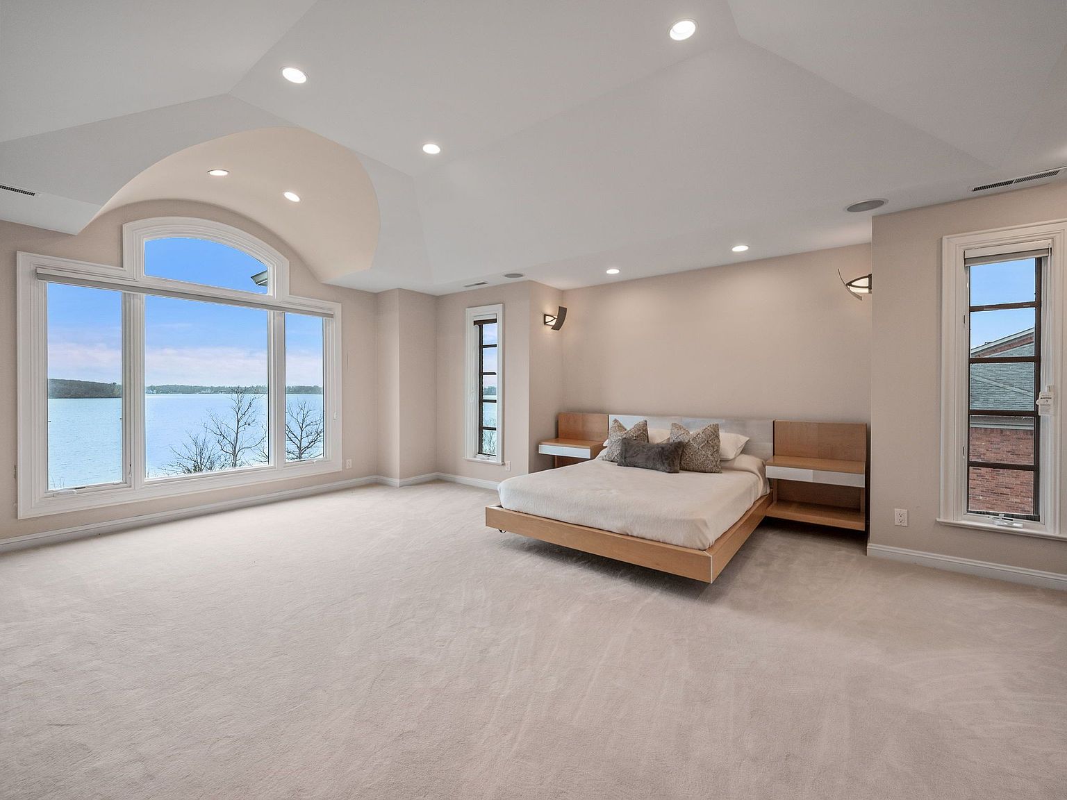 This is a spacious primary bedroom featuring a large window with a scenic lake view, a modern platform bed with built-in nightstands, and neutral-toned carpeting. The room is well-lit with recessed lighting and sconces, creating a serene and luxurious atmosphere. The color palette is soft and inviting, enhancing the room's tranquil ambiance.