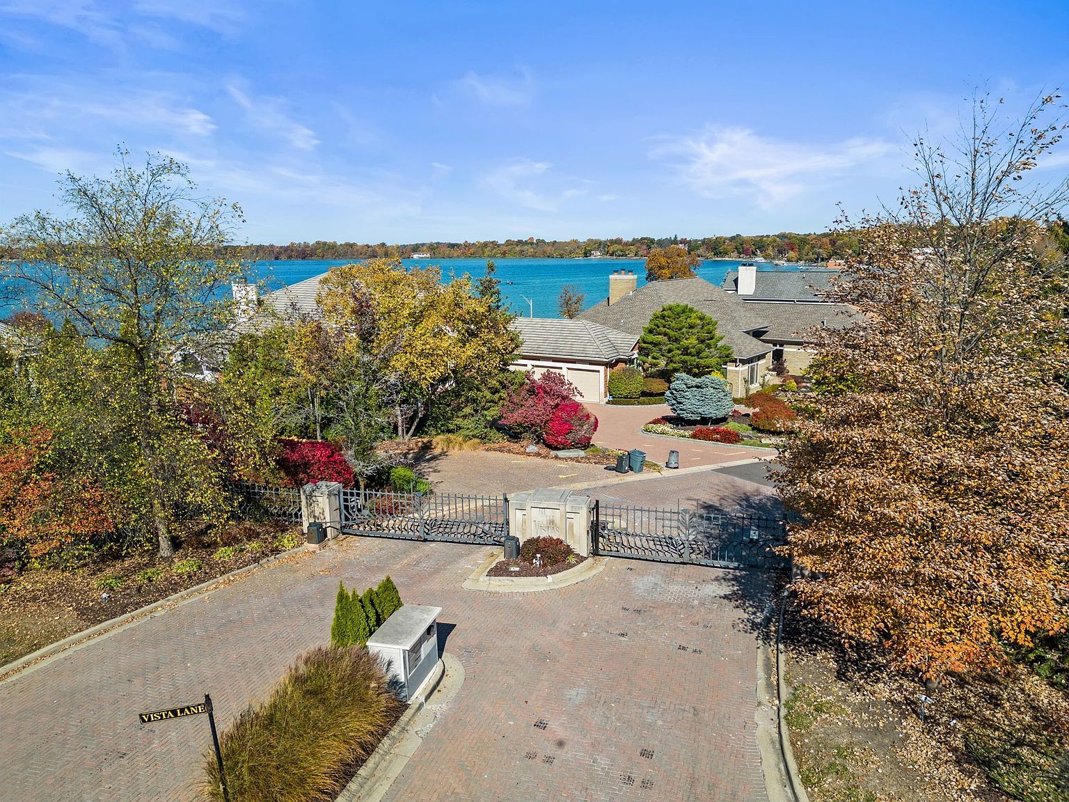This aerial shot showcases a luxurious residential entrance with a gated entry and meticulously maintained landscaping. The brick-paved driveway leads to upscale homes, with a serene lake visible in the background. Mature trees with autumn foliage add to the property's curb appeal and sense of exclusivity.