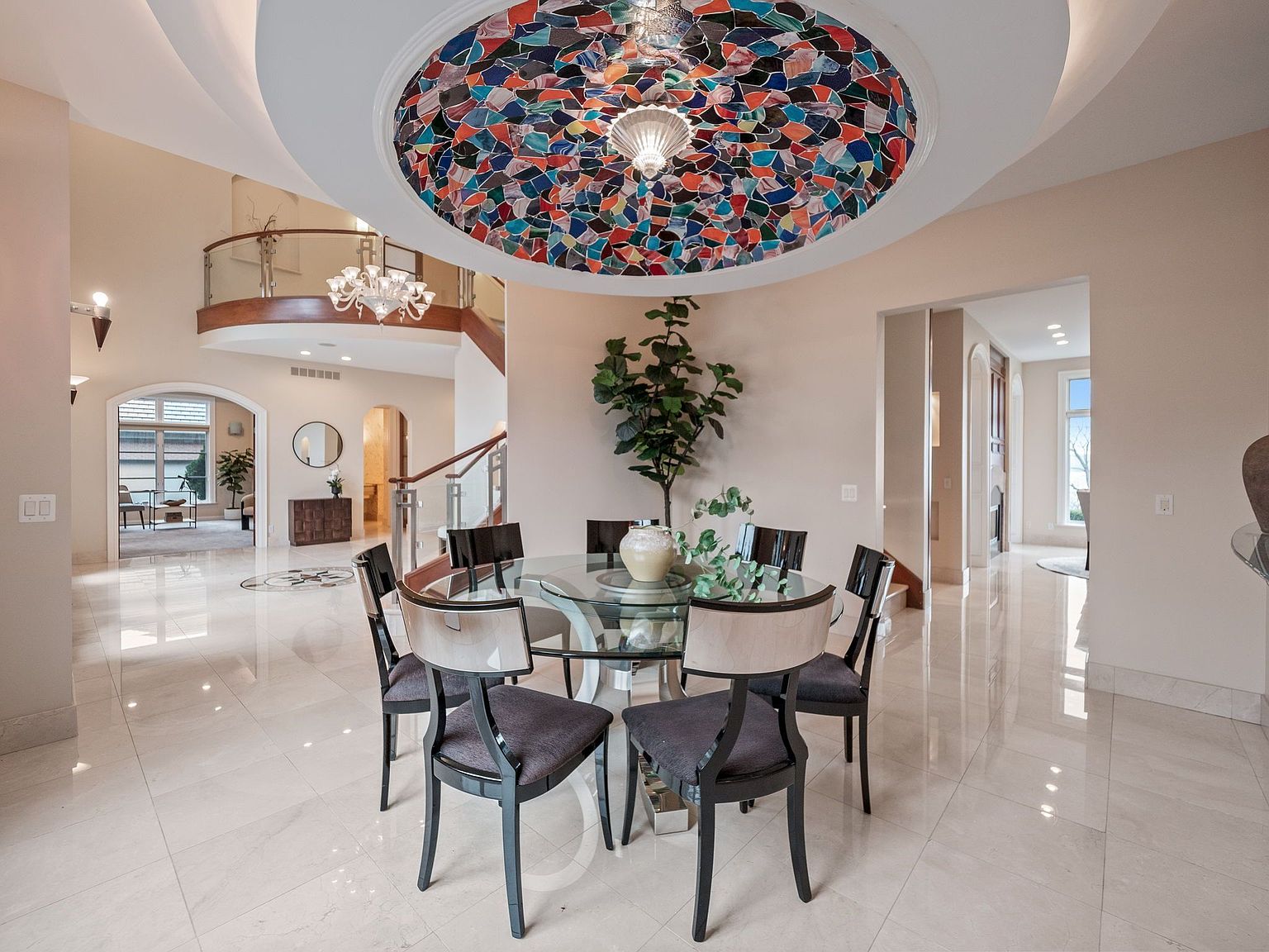 This interior shot showcases a luxurious dining area with a round glass table surrounded by modern chairs. A striking, colorful stained-glass dome ceiling feature adds a unique architectural element, complemented by a chandelier on the upper level. The polished marble flooring reflects light, enhancing the room's spacious and elegant ambiance.