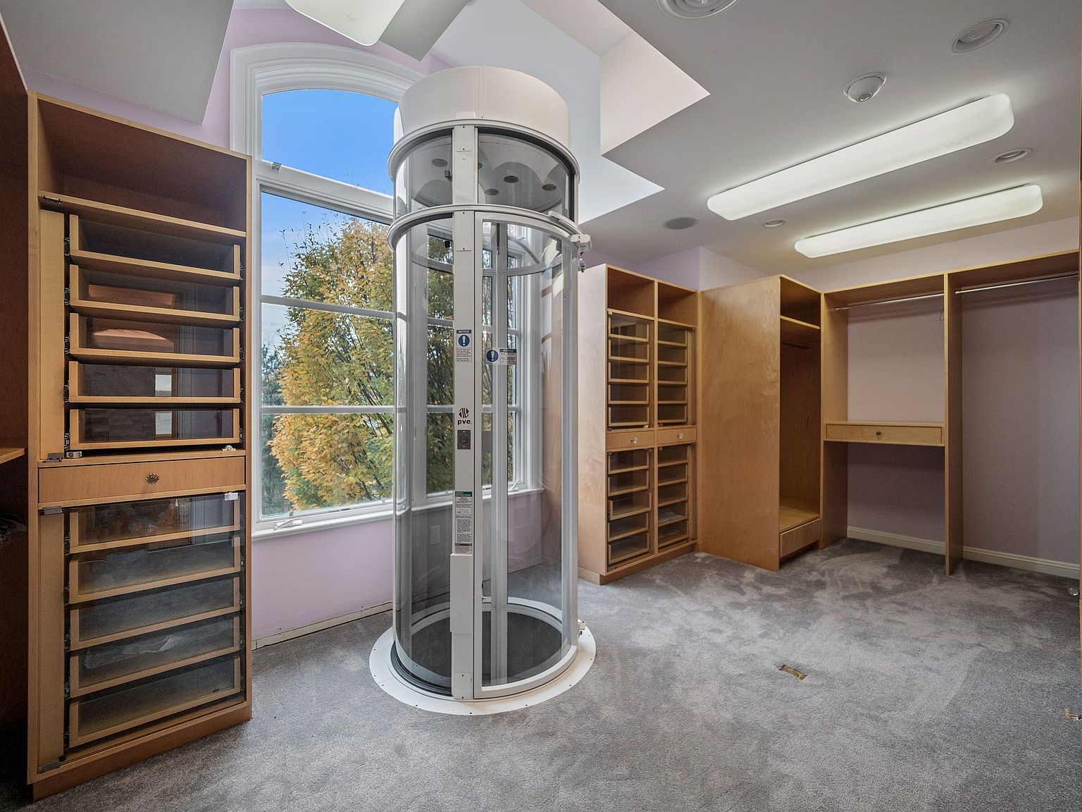 This is a spacious walk-in closet featuring custom wooden cabinetry and shelving, a unique cylindrical glass elevator, and soft gray carpeting. The closet is well-lit with recessed lighting and natural light from a large window, creating a luxurious and organized space. The presence of the elevator adds a high-end, accessible feature to the home.