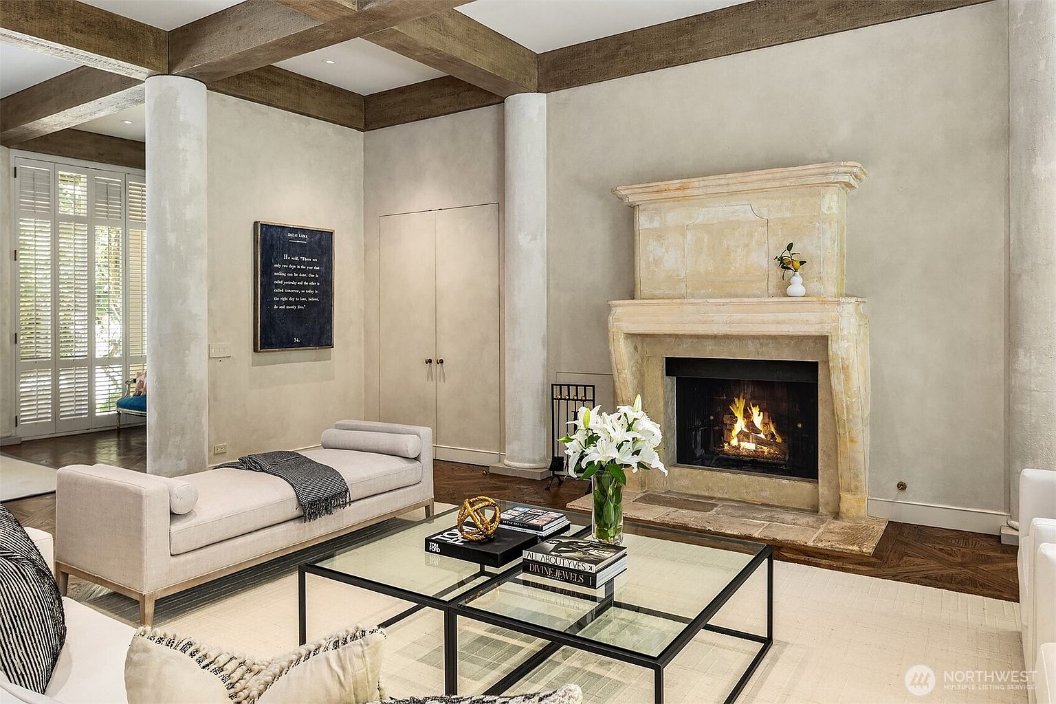 This is an interior shot of a living room featuring a stone fireplace with a burning fire, a glass-topped coffee table with books and decorative objects, and a neutral-toned daybed. The room has a sophisticated and elegant style, with exposed wooden beams on the ceiling and a large piece of artwork on the wall. The flooring is a mix of hardwood and a light-colored area rug.