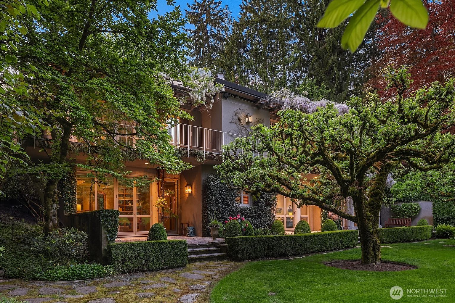 This is a beautifully landscaped front view of a two-story home. The house features a balcony with white railings, climbing wisteria, and large windows that suggest ample natural light. The manicured hedges, mature trees, and stone pathway create a welcoming and elegant curb appeal.