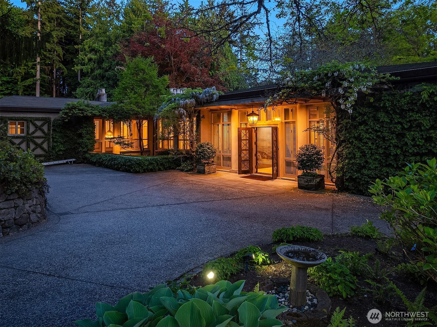 This image showcases the inviting entryway of a home, featuring a well-lit facade with warm lighting emanating from within. The entrance is framed by lush greenery and mature trees, creating a sense of privacy and tranquility. A paved driveway leads up to the house, and a birdbath adds a charming detail to the landscape.