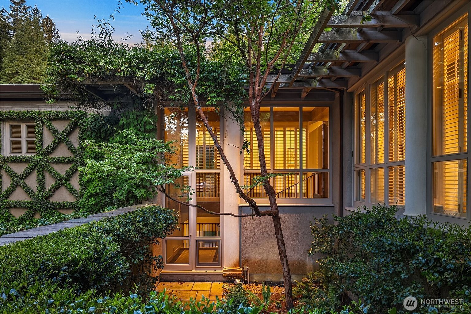 This image showcases a beautifully landscaped yard or garden area of a property. The exterior features a building with climbing vines and a trellis, creating a lush and inviting atmosphere. Large windows with blinds suggest a bright interior, while the surrounding greenery adds privacy and charm, enhancing the property's curb appeal.
