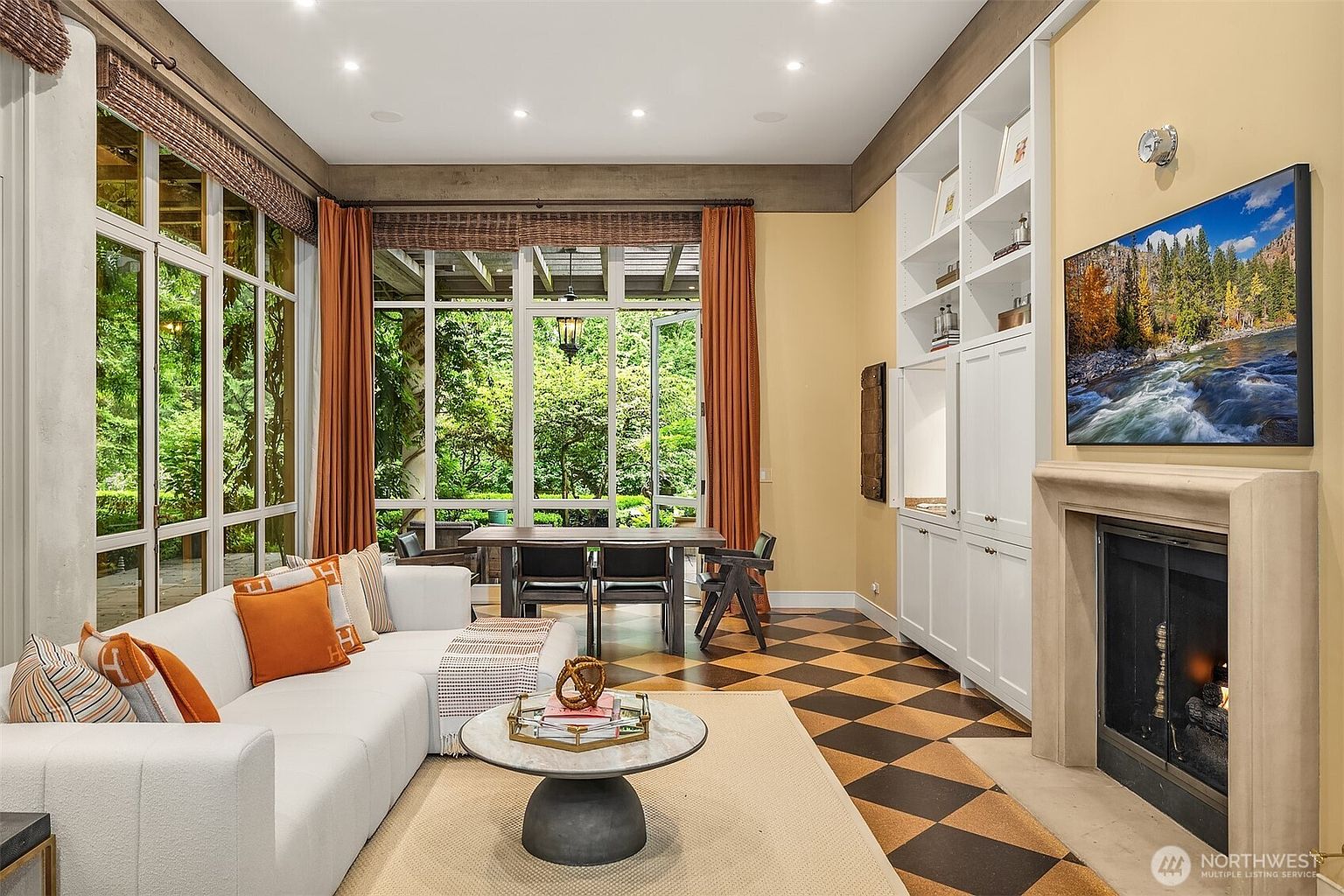 This is an interior shot of a living room featuring a large window with a view of a lush garden. The room is furnished with a white sectional sofa, a round coffee table, and a dining table with chairs. A fireplace with a television above it and built-in shelving add to the room's appeal, while the checkered floor adds a unique design element.