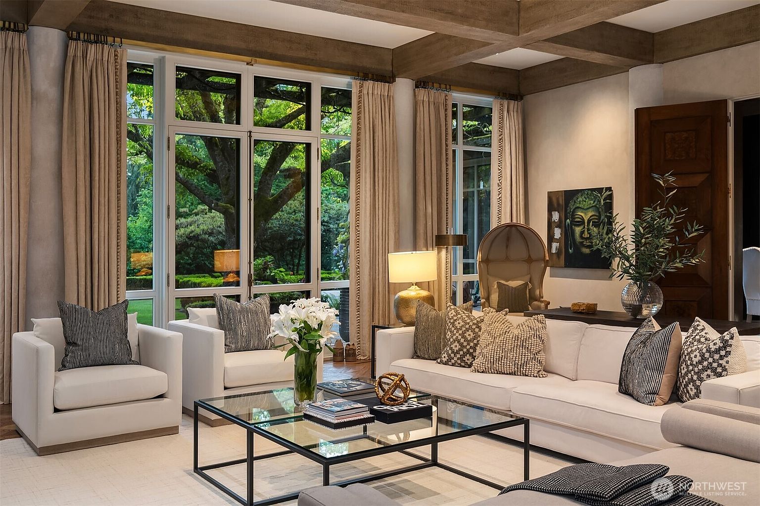 This is an interior shot of a luxurious living room, featuring a neutral color palette with white sofas and armchairs, complemented by patterned throw pillows. A glass coffee table sits in the center, and large windows offer a view of a lush garden. The room is well-lit and exudes a sense of sophisticated comfort.