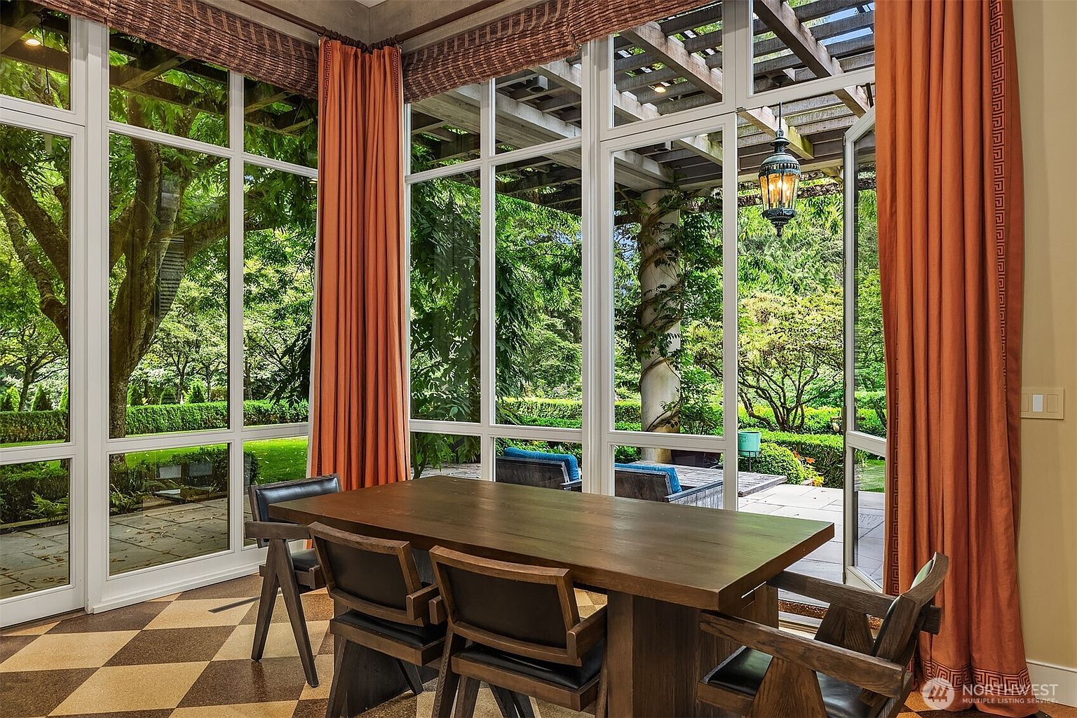 This interior shot showcases a dining room with a large wooden table and chairs, set against a backdrop of expansive windows offering views of a lush garden. The room features a checkered floor pattern and orange curtains, creating a warm and inviting atmosphere. The natural light floods the space, highlighting the connection between the interior and the exterior landscape.