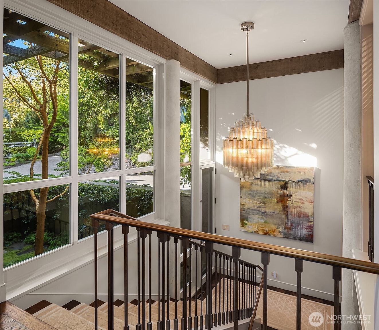 This interior shot showcases a well-lit hallway and staircase area. A large window provides ample natural light and a view of lush greenery outside. The staircase features a dark wood and carpet design, complemented by a modern metal railing. A striking chandelier hangs above, adding a touch of elegance to the space, while an abstract painting adorns the wall.