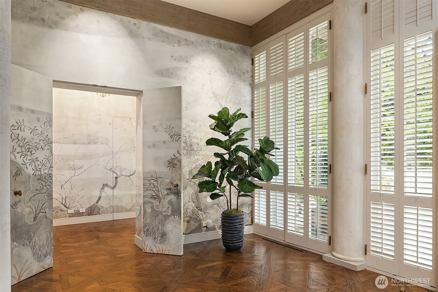 This interior shot showcases a hallway with unique architectural details, including decorative wall art and a parquet wood floor. The room features large shuttered windows that allow natural light to flood the space, complemented by a potted fiddle-leaf fig tree. The overall impression is one of sophisticated elegance and artistic flair.