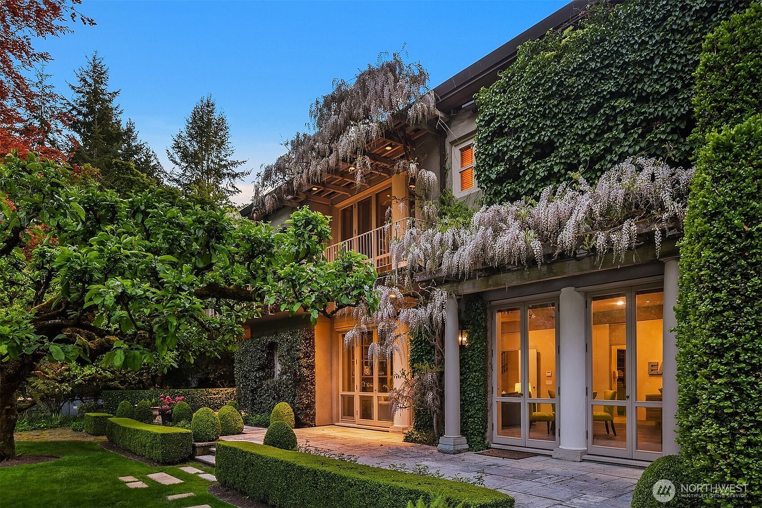 This image showcases a beautifully landscaped yard and garden area of a luxurious home. The exterior features lush greenery, including manicured hedges, mature trees, and climbing wisteria adorning the building's facade. Large glass doors provide access to the interior, while a stone patio offers an inviting outdoor space.