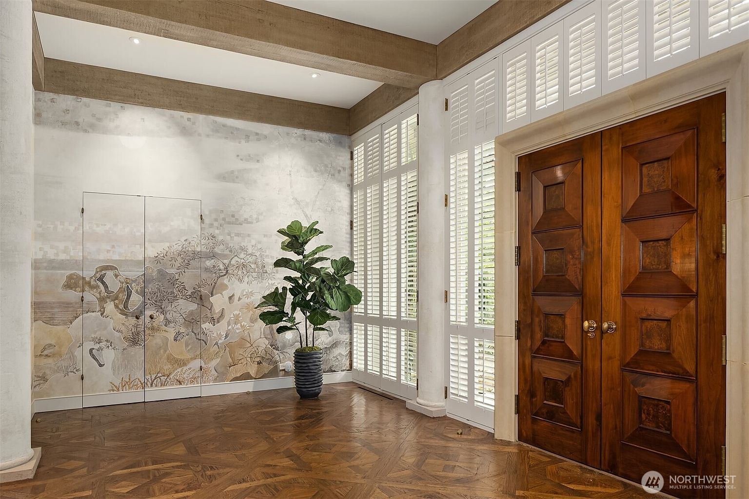 This interior shot showcases a grand hallway featuring a herringbone wood floor, a decorative wall mural, and a large potted plant. The space is illuminated by natural light filtering through shuttered windows, and a set of ornate wooden double doors adds a touch of elegance. The overall impression is one of sophistication and classic design.