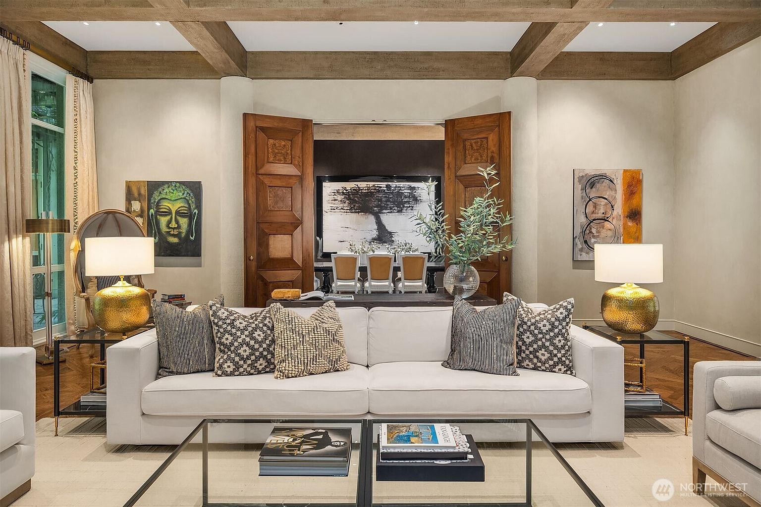 This is an interior shot of a luxurious living room, featuring a white sofa adorned with decorative pillows, a glass coffee table with books, and side tables with golden lamps. The room is decorated with artwork and has wooden ceiling beams, creating a sophisticated and inviting atmosphere. The open wooden doors lead to another room, adding depth and interest to the space.