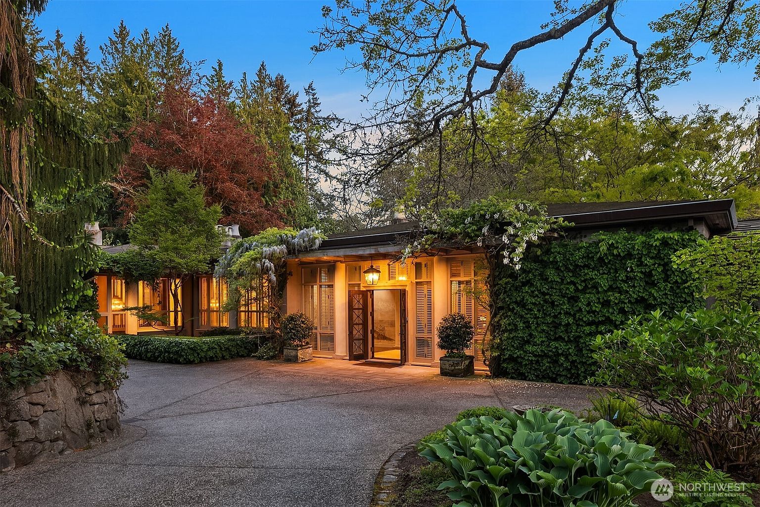 The image showcases the grand entryway of a home, framed by lush greenery and mature trees. The entrance features a partially open, ornate wooden door flanked by shuttered windows, creating a welcoming and sophisticated ambiance. A paved driveway leads up to the house, enhancing the property's curb appeal and suggesting a private, well-maintained estate.