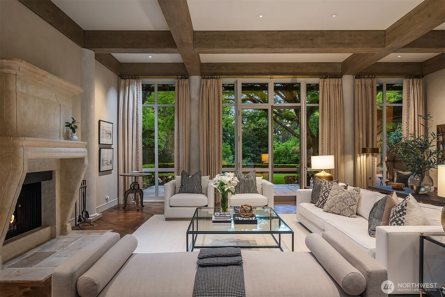 This is an interior shot of a luxurious living room featuring a stone fireplace, exposed wooden beams, and large windows overlooking a lush garden. The room is furnished with white sofas, armchairs, and a glass coffee table, creating a bright and inviting space. The perspective is from the front of the room, showcasing the depth and connection to the outdoor scenery.