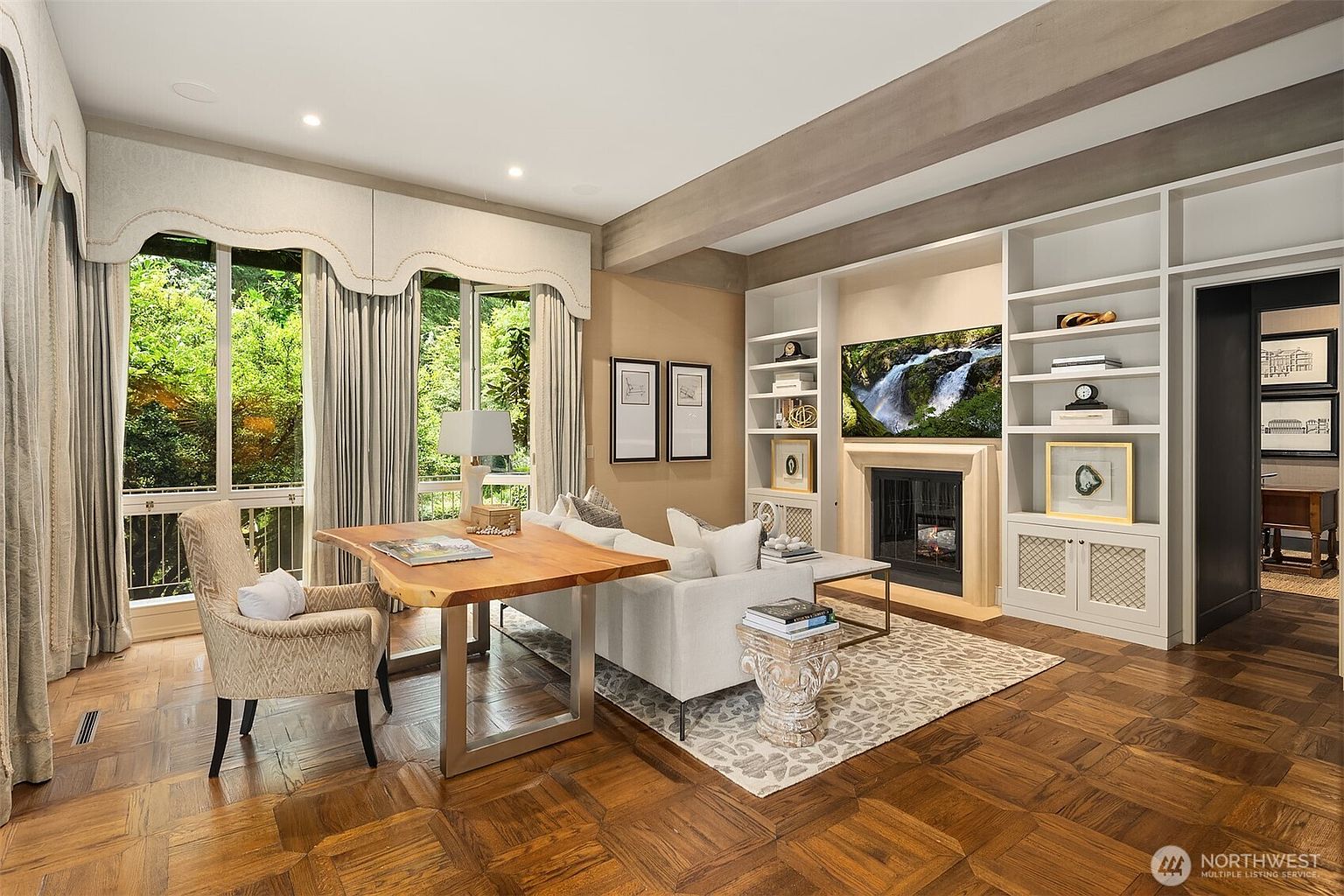 This is an interior shot of a living room featuring a wooden parquet floor, a light-colored sofa, and a wooden desk with a chair. The room has large windows with elegant curtains, providing natural light and a view of the greenery outside. A fireplace with a TV above it is flanked by built-in bookshelves, adding to the room's cozy and sophisticated ambiance.