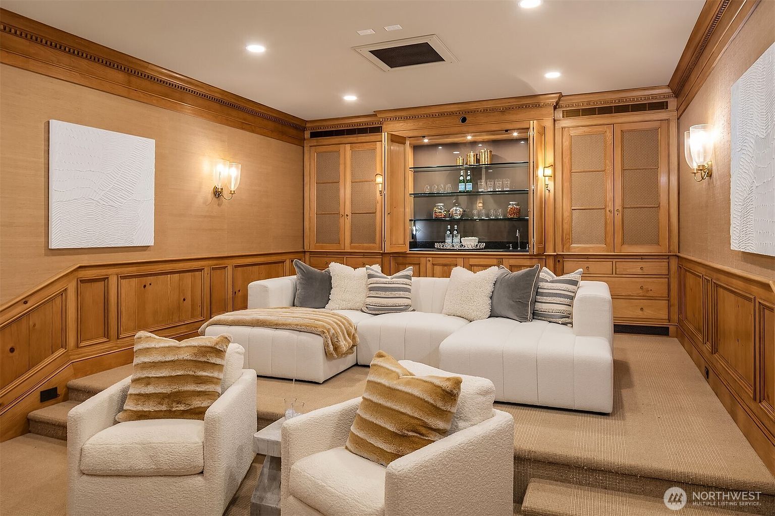 This is an inviting living room featuring a large, white sectional sofa with decorative pillows, complemented by two armchairs with striped cushions. The room is characterized by wood paneling and built-in cabinetry, including a bar area with glass shelves. The warm lighting and neutral color palette create a cozy and sophisticated atmosphere.
