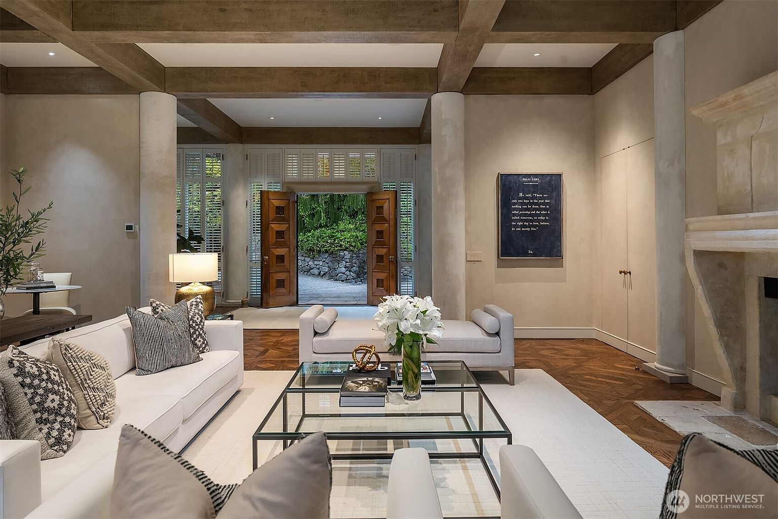 This is an interior shot of a luxurious living room, featuring a neutral color palette with white sofas and a glass coffee table. The room is characterized by its exposed wooden beams, columns, and hardwood floors, creating a sophisticated and inviting atmosphere. A large doorway leads to an outdoor area, enhancing the sense of space and connection to nature.