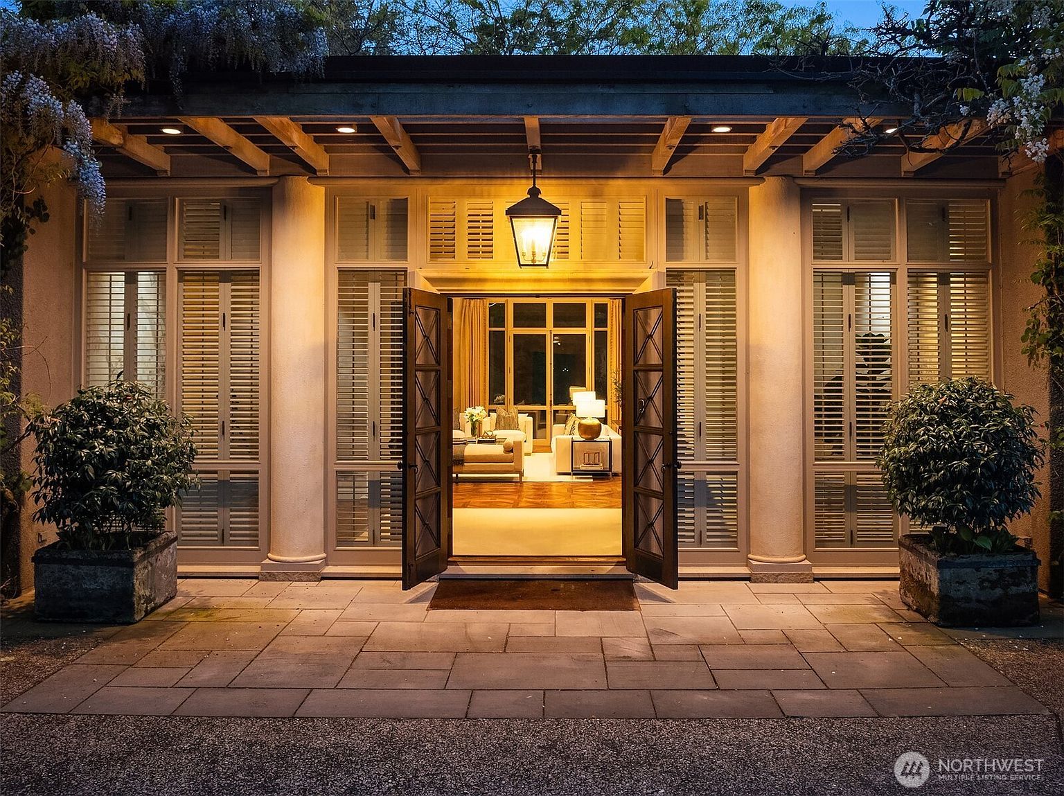 This image showcases a grand entryway with double doors leading into a well-lit living space. The entrance is framed by large windows with shutters and supported by elegant columns, creating a symmetrical and inviting facade. A hanging lantern illuminates the doorway, and potted plants add a touch of greenery to the stone-paved entrance, enhancing the property's curb appeal.