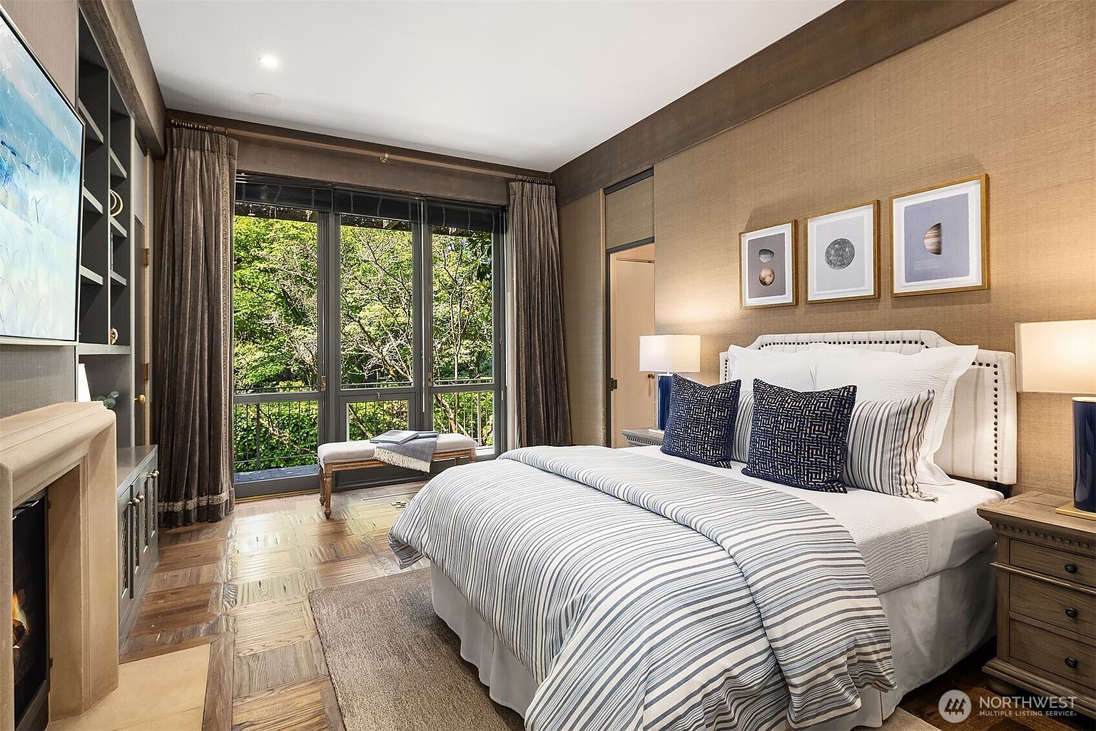This is a primary bedroom featuring a large bed with striped bedding and decorative pillows, flanked by nightstands and lamps. The room has a large window overlooking lush greenery, providing natural light and a sense of tranquility. The walls are textured, and artwork hangs above the bed, contributing to a sophisticated and comfortable atmosphere.