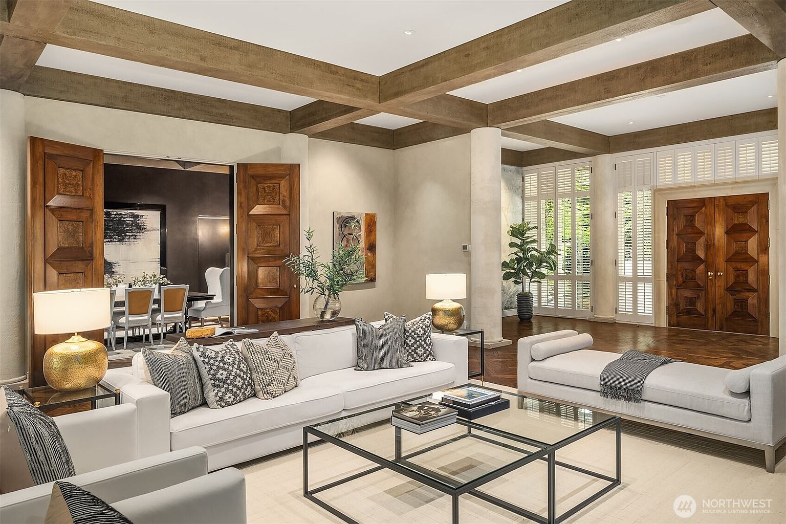 This is an interior shot of a luxurious living room featuring a coffered ceiling with wooden beams, a large white sofa adorned with decorative pillows, and a glass-topped coffee table. The room is well-lit, showcasing hardwood floors and elegant wooden doors, creating a warm and inviting atmosphere. A daybed sits to the right of the sofa, and the room opens into a dining area.