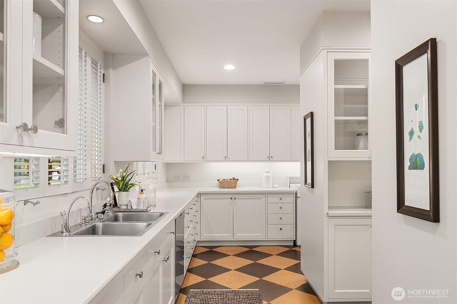 This is a well-organized laundry room featuring white cabinetry, a stainless steel sink, and a checkerboard-patterned floor. The room is brightly lit with recessed lighting and under-cabinet lights, creating a clean and functional space. A framed artwork adds a touch of elegance to the room.
