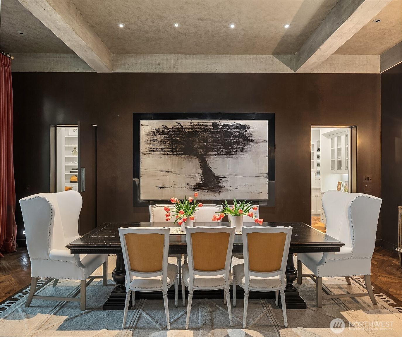 This is an interior shot of a dining room featuring a dark wood table surrounded by white upholstered chairs with light wood accents. A large abstract painting hangs on the wall, and the room is illuminated by recessed lighting in the coffered ceiling. The overall impression is elegant and sophisticated.