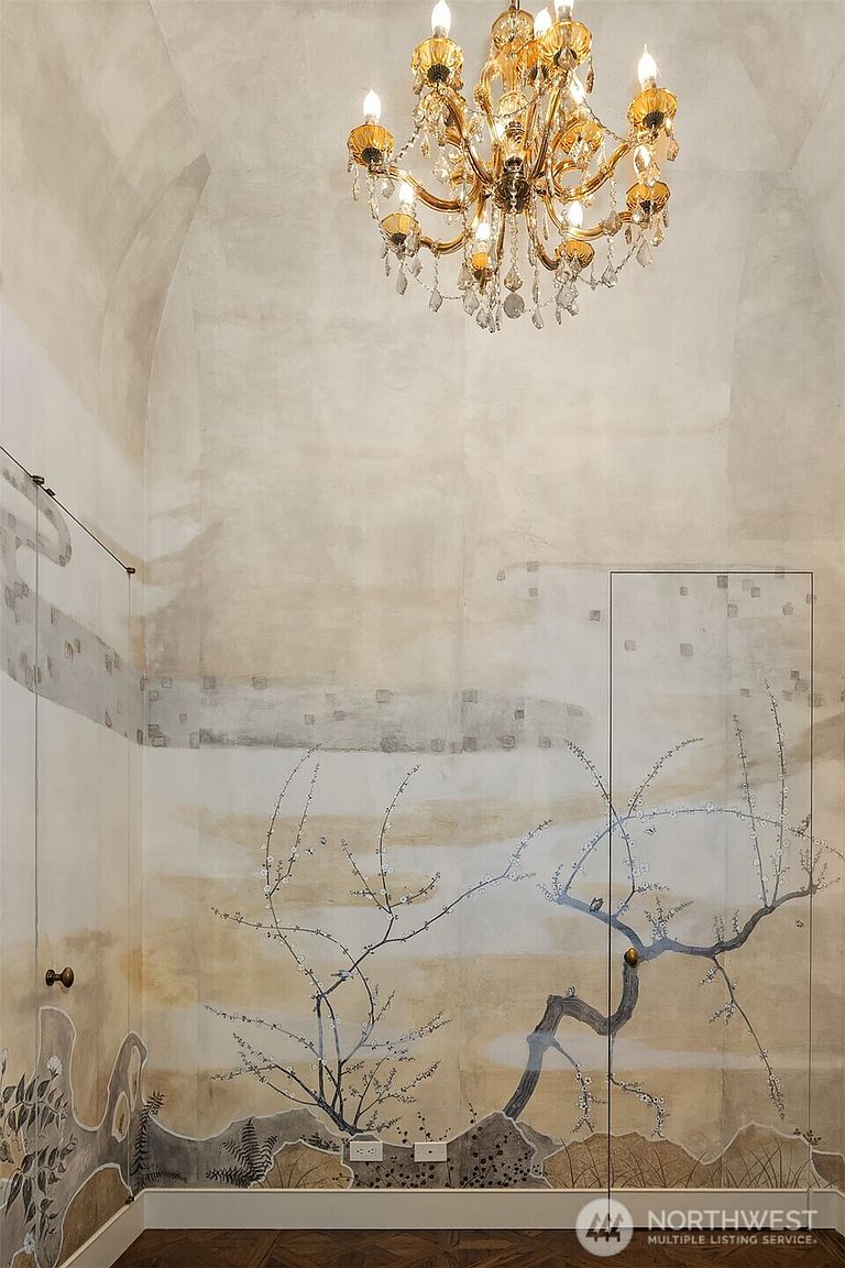 This interior shot showcases a hallway or stairwell area with unique, artistic wallpaper featuring a tree motif and a neutral color palette. A gold chandelier hangs from the ceiling, adding a touch of elegance to the space. The flooring appears to be hardwood, contributing to the room's sophisticated aesthetic.