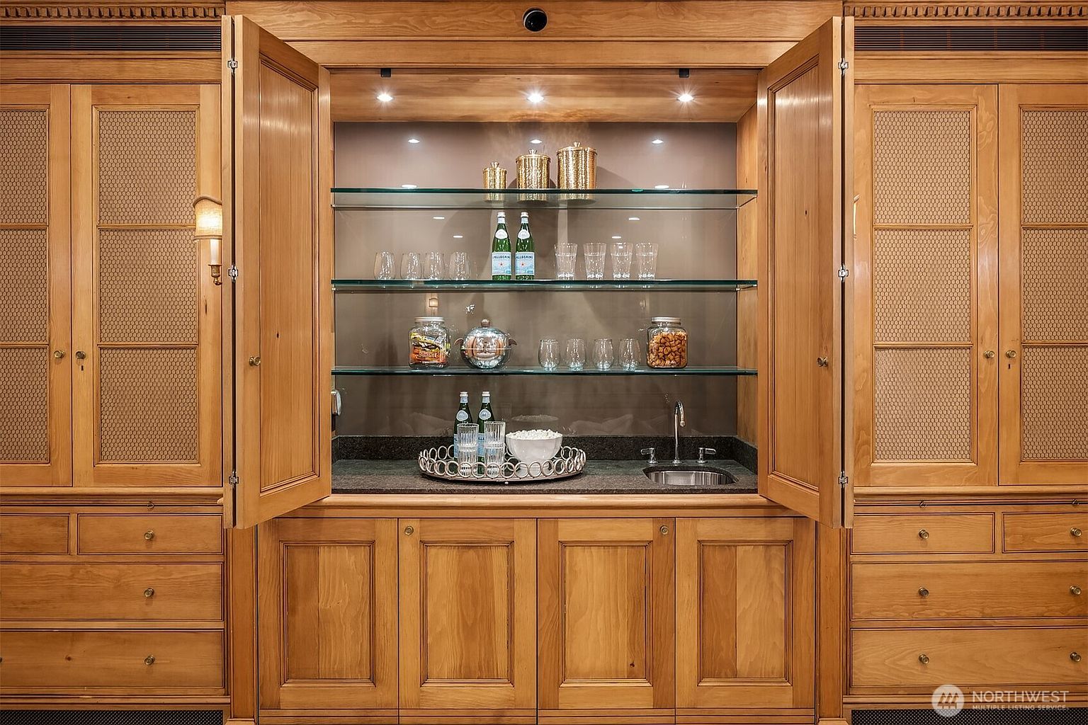 This image showcases a well-organized and elegant pantry. The pantry features wooden cabinetry with mesh door inserts on the sides, glass shelving, and built-in lighting. The shelves are stocked with glassware, bottles, and decorative jars, creating a sophisticated and functional storage space.