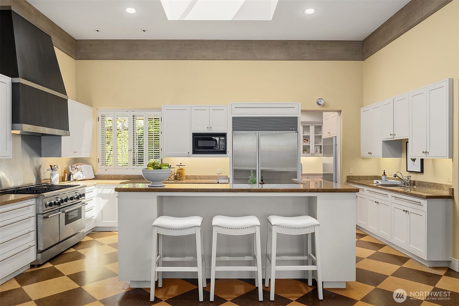 This is a well-lit kitchen with a high ceiling and a skylight. The kitchen features white cabinetry, stainless steel appliances, and a large island with three white bar stools. The floor has a checkered pattern, and the walls are painted in a light yellow tone, creating a warm and inviting atmosphere.