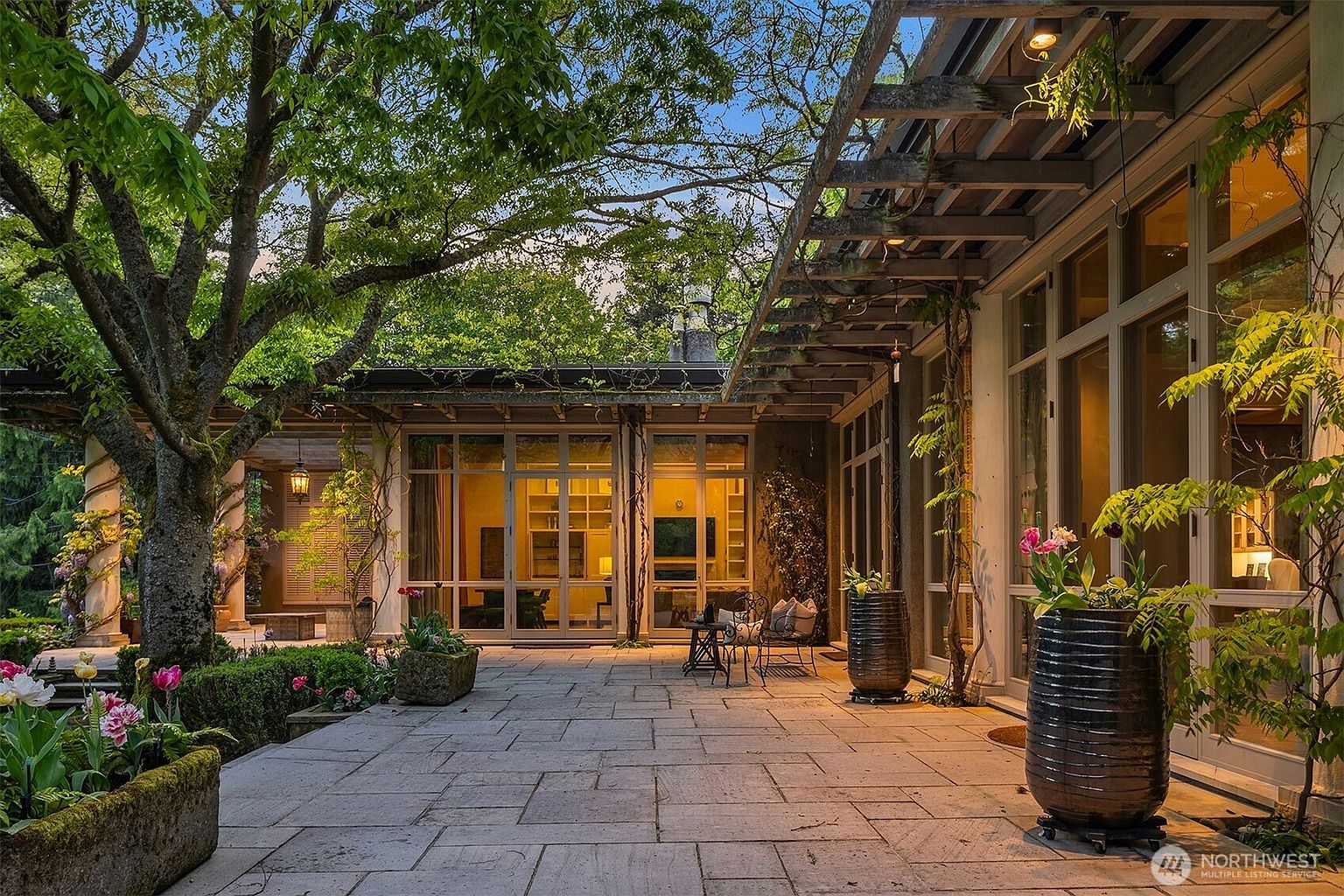 This image showcases a beautifully designed patio area, featuring a stone-paved surface that extends from the building's large windows and doors. The patio is adorned with potted plants and flowers, creating a serene outdoor living space. A wooden pergola provides partial shade, while mature trees offer additional greenery and privacy, enhancing the property's appeal for relaxation and entertainment.