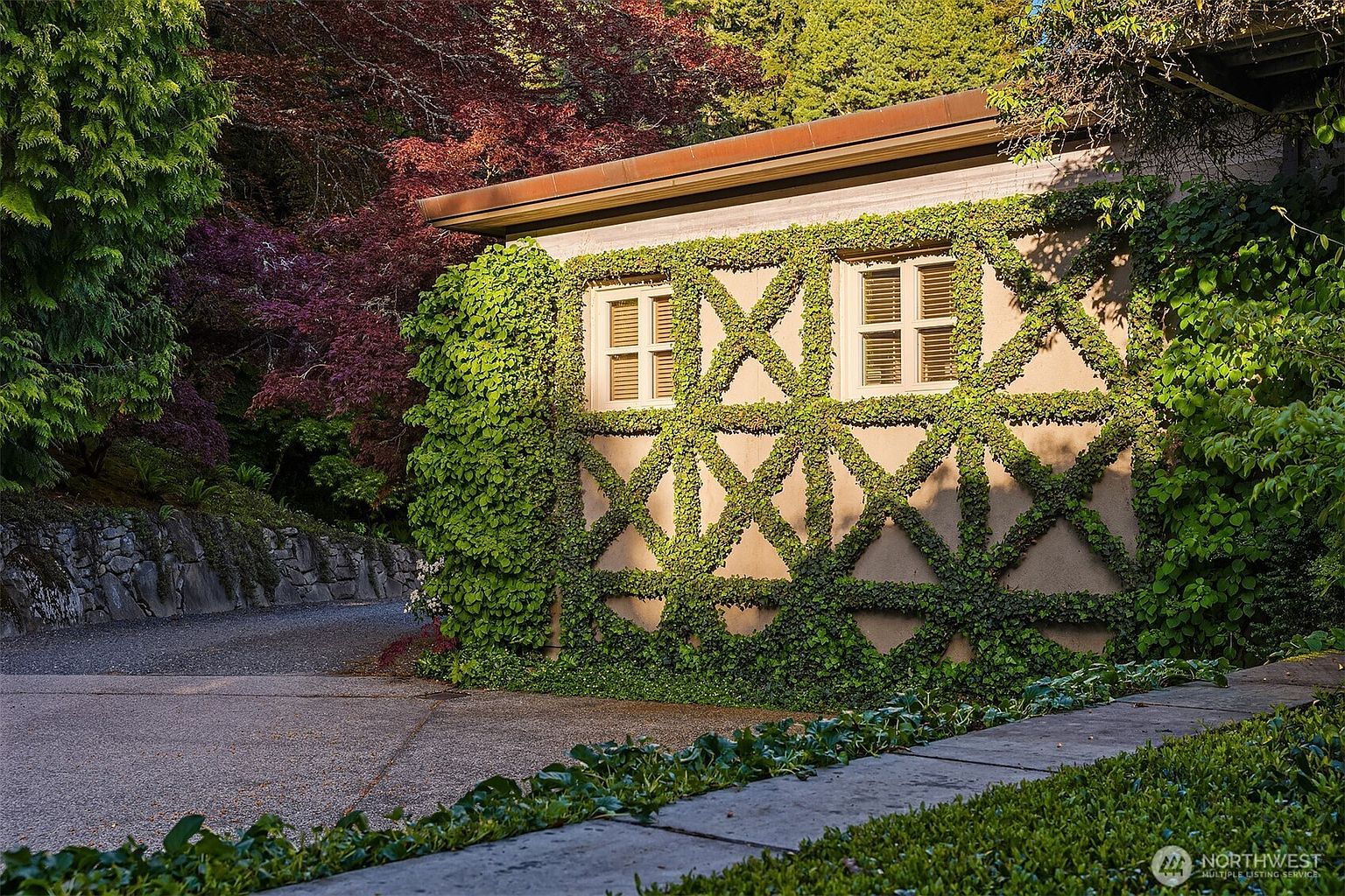 This is a side view of a building with a unique exterior design. The wall is covered in a diamond-patterned trellis with climbing ivy, creating a visually appealing and natural aesthetic. Two windows with light-colored frames and blinds are visible, adding to the charm of the building's facade.