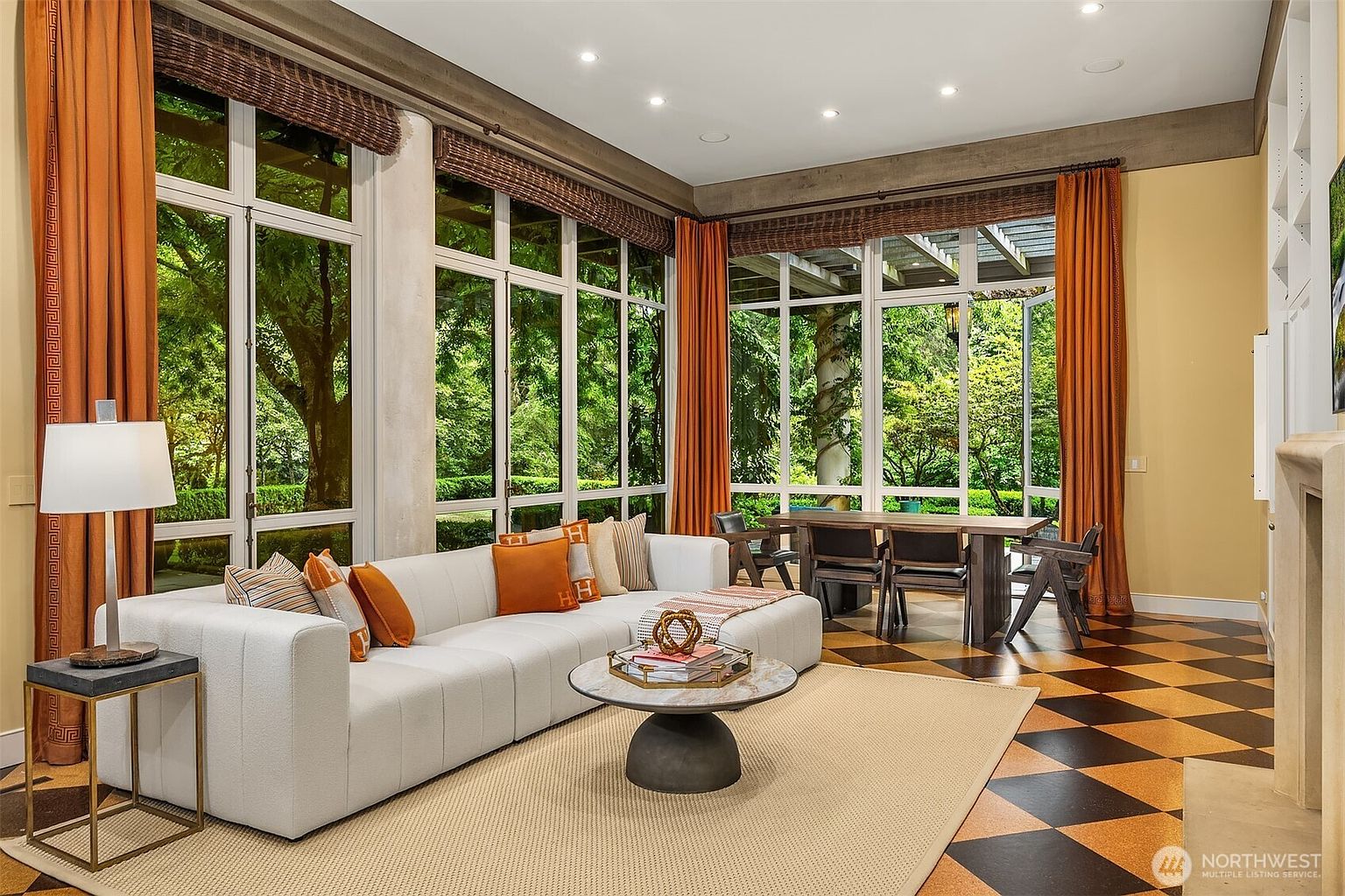 This is an interior shot of a living room featuring a large white sectional sofa with orange accent pillows, a round coffee table, and a side table with a lamp. The room is bright and airy, with large windows offering views of a lush garden. The flooring is a checkered pattern, and the overall style is modern and sophisticated.