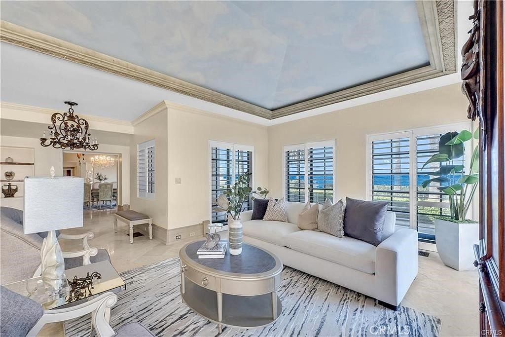 This elegant living room features a distinctive ceiling painted with a cloud mural, framed by decorative molding. The space is furnished with a plush white sofa, a central oval coffee table, and a patterned area rug, all oriented toward large windows that offer a view of the ocean. A crystal chandelier hangs in the adjacent dining area, contributing to the sophisticated and airy atmosphere of the home.