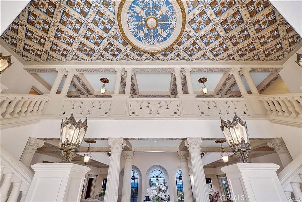 This grand foyer features a dramatic double staircase with ornate balustrades and a central landing overlooking the lower level. The ceiling is a focal point, showcasing an intricate, multi-colored coffered design with a central medallion and decorative molding. Large, ornate lanterns provide lighting, while classical columns and arched windows contribute to the opulent, palatial aesthetic of the space.