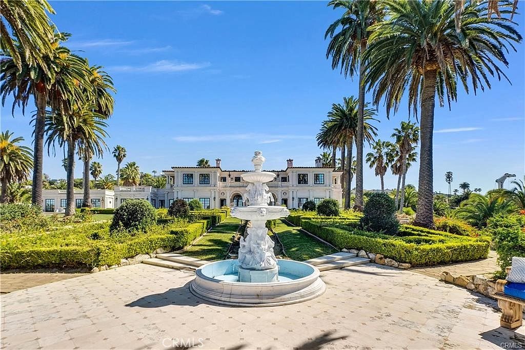 This grand estate features a meticulously manicured formal garden centered around an ornate, multi-tiered stone fountain. The symmetrical landscape design includes neatly trimmed hedges and tall, mature palm trees that frame the impressive white mansion in the background. The perspective is a wide, eye-level shot capturing the expansive stone-paved courtyard and the elegant architectural details of the residence.