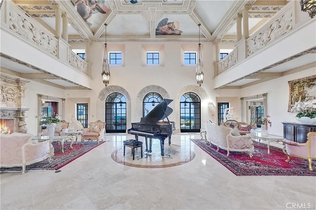 This grand, double-height living room features an opulent, classical design with a central grand piano positioned on a circular stone inlay. The space is framed by three arched glass doors, ornate ceiling murals, and symmetrical balconies overlooking the main floor. Elegant seating areas with plush furniture and intricate patterned rugs are arranged on either side, creating a sophisticated and symmetrical atmosphere.