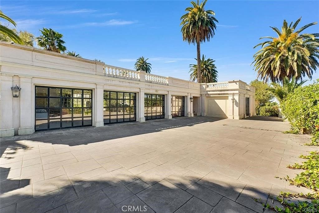 This image captures a spacious, paved motor court featuring a multi-bay garage with large, grid-patterned glass doors. The structure is designed in a classical architectural style with light-colored masonry, decorative balustrades along the roofline, and symmetrical detailing. Tall palm trees frame the scene against a clear blue sky, creating an elegant and expansive outdoor arrival area.