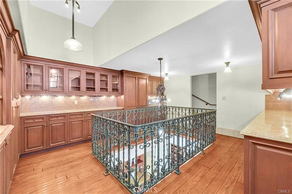 This interior view captures an upper-level landing area featuring extensive custom wood cabinetry and a decorative wrought-iron railing overlooking the space below. The area is illuminated by a prominent hanging chandelier and recessed lighting, with warm-toned wood flooring throughout. The perspective looks across the landing toward a staircase, highlighting the open, airy feel created by the high ceilings.