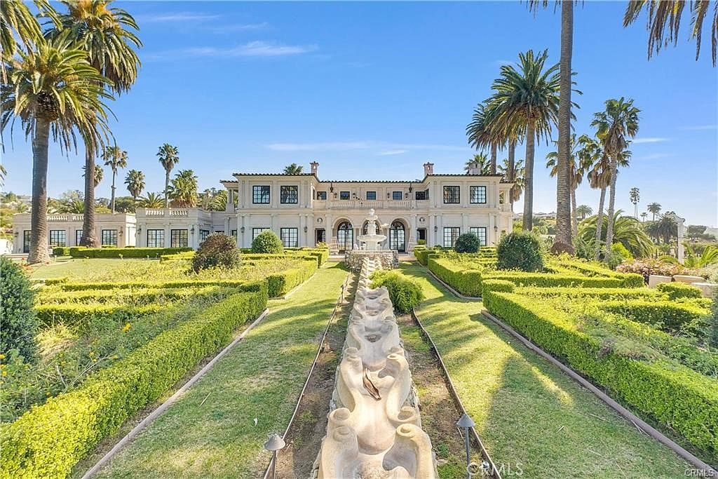 This grand Mediterranean-style estate is captured from a low-angle perspective, emphasizing the symmetrical architecture and the formal garden path leading to the residence. A long, tiered stone water feature serves as a central axis, flanked by manicured hedges and lush green lawns. Tall palm trees frame the structure, which features a light-colored facade, multiple arched windows, and a classic balcony, creating an atmosphere of opulence and refined elegance.