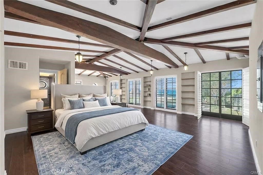 This spacious primary bedroom features a vaulted ceiling with exposed dark wood beams and dark hardwood flooring throughout. A large, light-colored upholstered bed serves as the focal point, complemented by a blue patterned area rug and matching bedside tables. The room is brightened by multiple pendant lights and large glass doors that provide a view of the outdoor greenery.