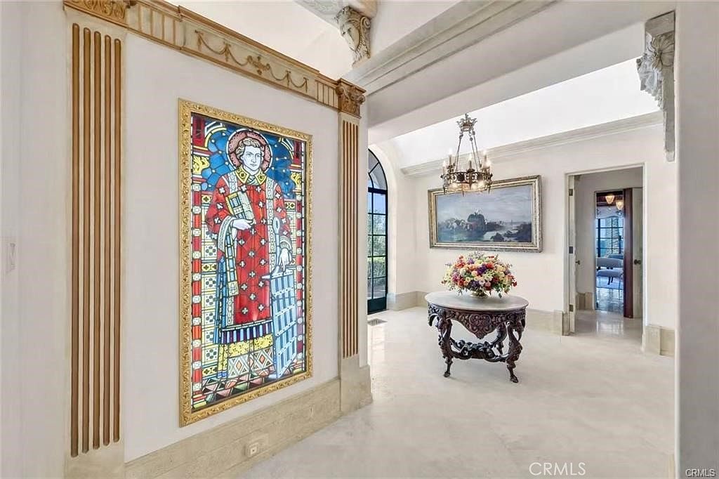 This elegant foyer or hallway features a striking stained-glass panel depicting a religious figure, framed by decorative columns and molding. The space is illuminated by a classic chandelier hanging above a round pedestal table adorned with a vibrant floral arrangement. The perspective looks through an arched opening into a bright, secondary room, highlighting the home's sophisticated architectural details and polished light-colored flooring.