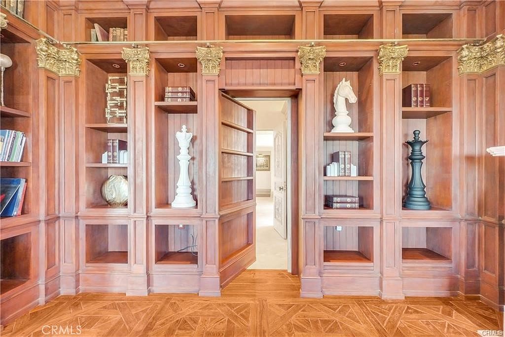 This sophisticated study features extensive custom wood cabinetry with intricate molding and decorative gold-toned capitals. A hidden door integrated into the shelving unit reveals a glimpse into an adjacent room, adding a sense of mystery and architectural interest. The space is accented with large decorative chess pieces and books, all set against a rich, warm-toned wood floor with a geometric parquet pattern.