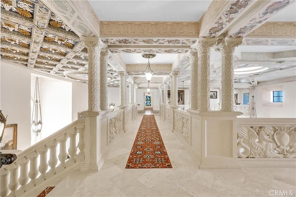 This grand, opulent hallway features a long, ornate runner rug centered on light-toned stone flooring, flanked by a series of classical columns and intricate balustrades. The ceiling is highly decorative, showcasing elaborate coffered detailing and painted murals that extend down the upper walls. The perspective is a symmetrical, eye-level shot looking down the length of the gallery, emphasizing the architectural scale and luxurious, old-world aesthetic.