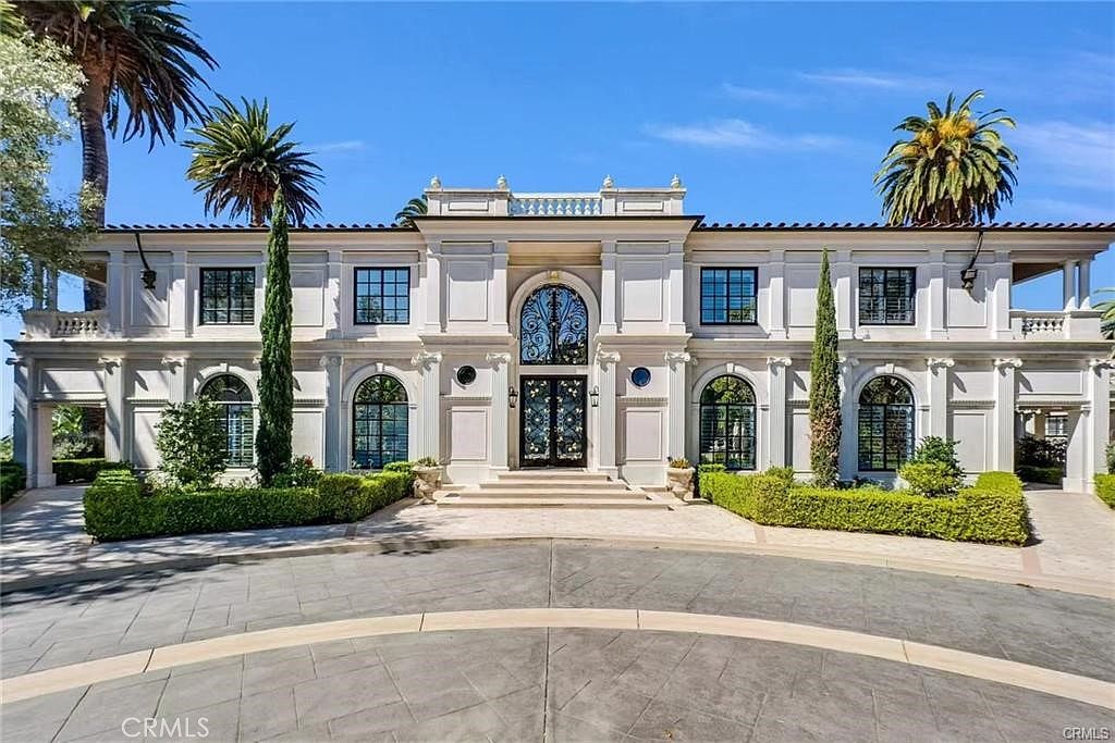 This grand, symmetrical estate features a stately neoclassical facade with a prominent arched entryway and ornate metal double doors. The exterior is finished in a light-toned stucco, accented by tall, multi-pane windows and manicured hedges that frame the paved circular driveway. Two tall, slender cypress trees stand as focal points on either side of the entrance, enhancing the formal and imposing architectural presence of the home.