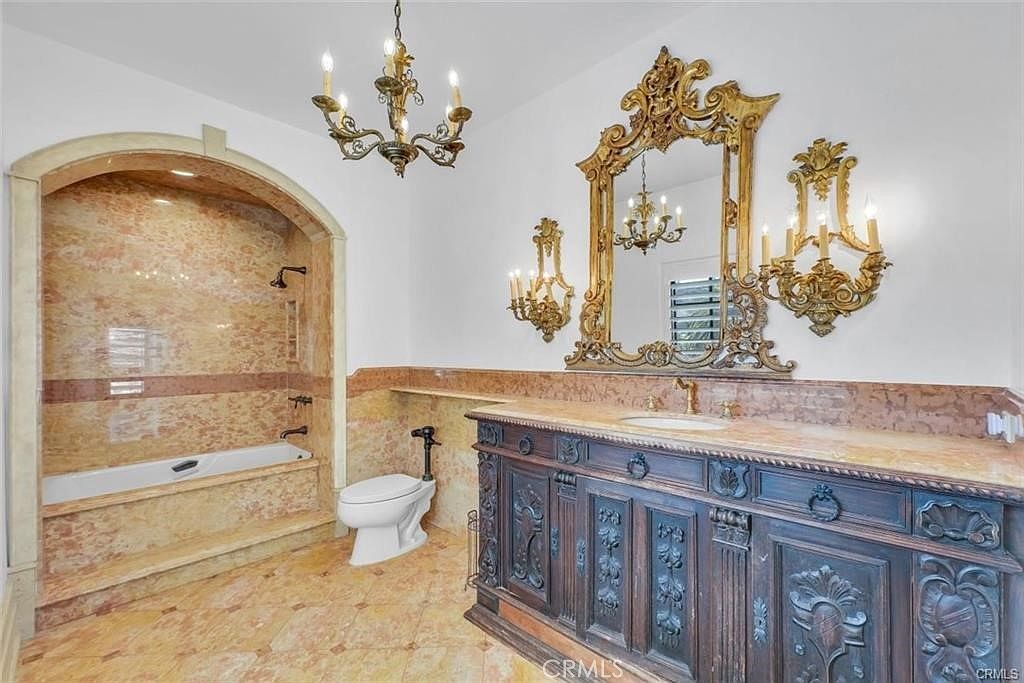 This opulent primary bathroom features extensive use of patterned stone on the walls and flooring, creating a cohesive and luxurious aesthetic. The space includes a built-in bathtub set within an arched alcove, a dark wood vanity with an ornate mirror, and decorative wall sconces that provide warm lighting. The perspective is a wide-angle shot capturing the entire room, emphasizing the dramatic, traditional design elements.