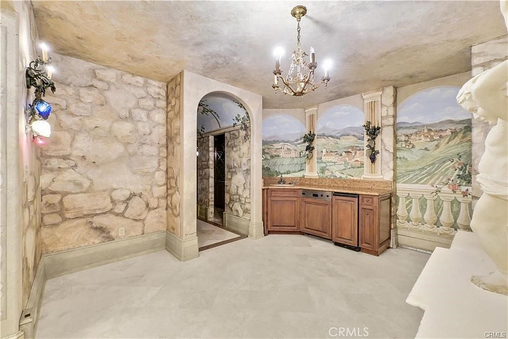 This unique interior space features a Mediterranean-inspired design with faux-stone walls and a hand-painted mural depicting a rolling landscape. A built-in wet bar with wood cabinetry and a small refrigerator sits beneath the mural, while an ornate chandelier hangs from the textured ceiling. The room is framed by an arched stone doorway leading to an adjacent area, creating an immersive, old-world atmosphere.
